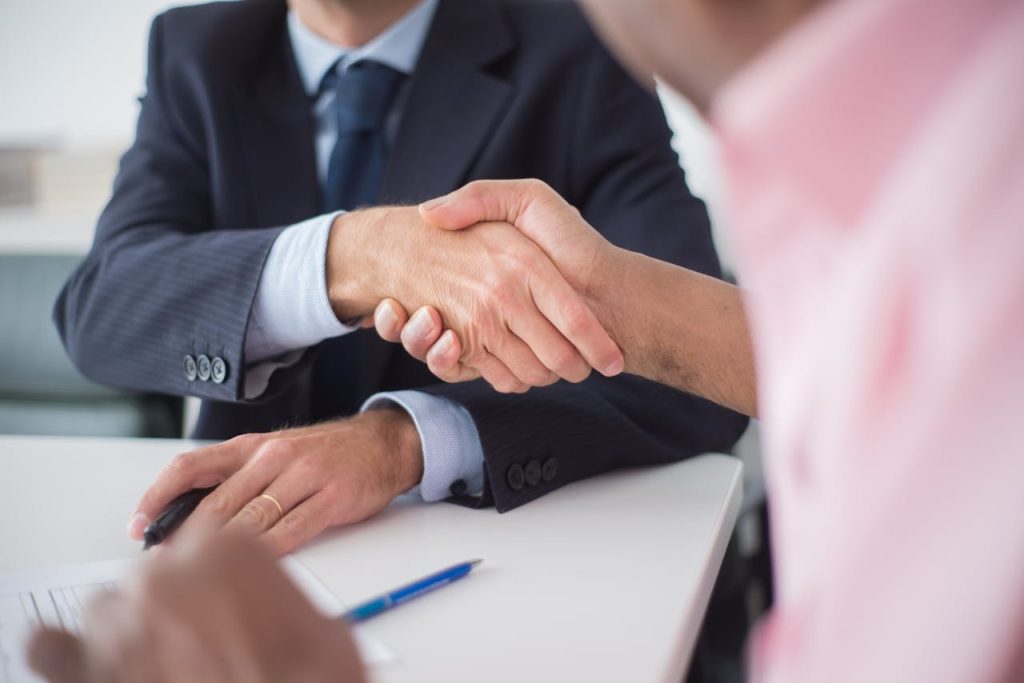 close-up-of-shaking-hands-between-men-8441790 Two businessmen shaking hands across table, symbolizing agreement and partnership in an office environment.