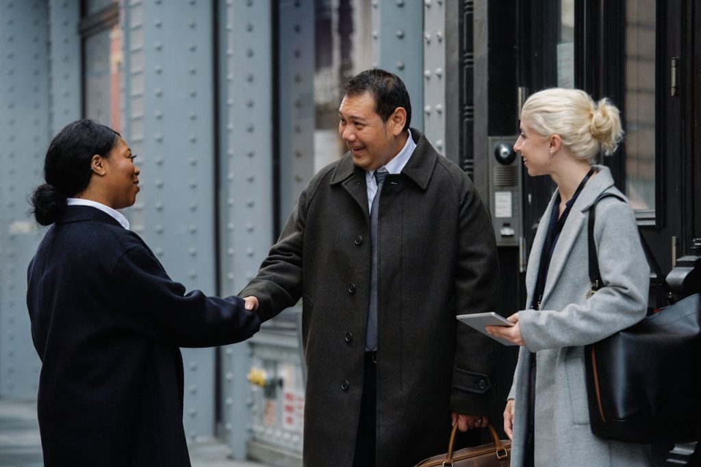 smiling-diverse-coworkers-shaking-hands-on-street-before-meeting-5668461 A diverse group of professionals meeting and greeting with a handshake outdoors.